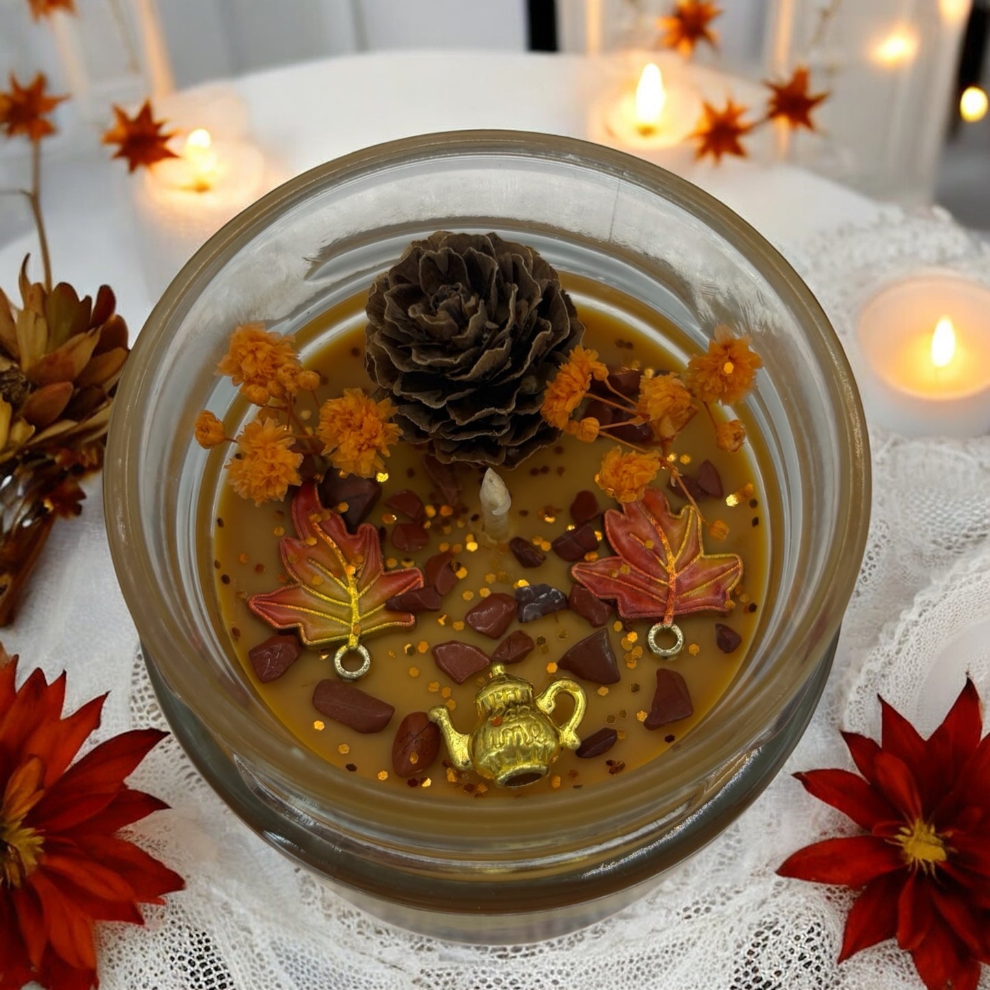Decorative autumn candle featuring pine cone, orange flowers, and leaf ornaments, surrounded by warm fall colors.